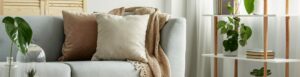 Divano grigio chiaro con cuscini neutri e coperta intrecciata, accanto a scaffale con piante verdi e libri in un soggiorno luminoso e moderno.